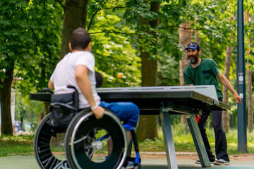 Inclusiveness A disabled man in a wheelchair plays ping pong with an older man in a city park...