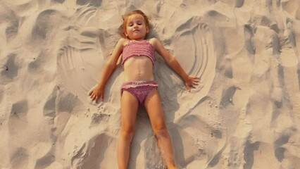 A child playing in a brown swimsuit lying on the beach on the sand makes an snow angel - Powered by Adobe