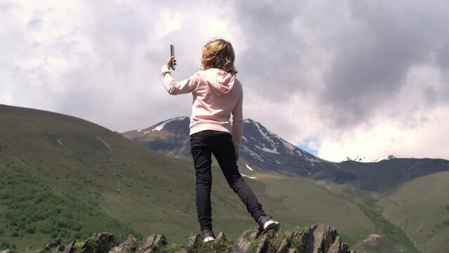 Little girl taking photos of mountains while standing on the edge of cliff, slow motion. Adventure and travel concept