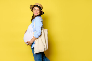 Authentic pregnant woman holds her belly, smiles cutely looking aside at copy space, carrying eco linen shopping bag