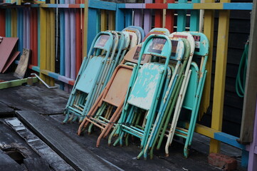 Rusty painted folded metal chairs with paint peeling off leaning on colourful fence in a harbour with wooden floor in a outdoor environment. Wet wooden floor in an outdoor environment.