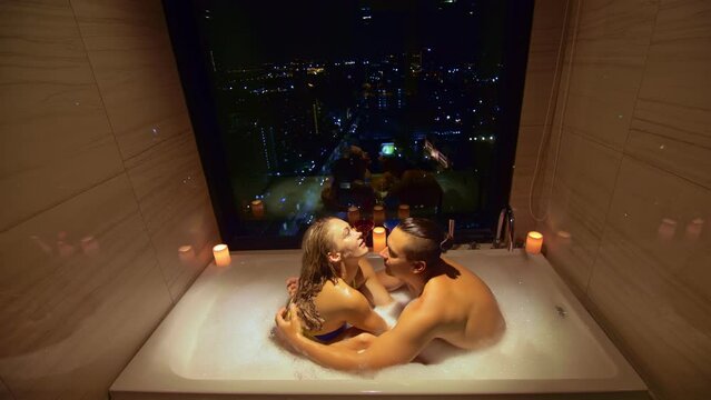 Love couple enjoys romantic honeymoon in hotel. Honeymoon vacation romantic atmosphere in candlelight. Young married couple relax in bathtub with window view of city night.Embracing in foam, kiss hug