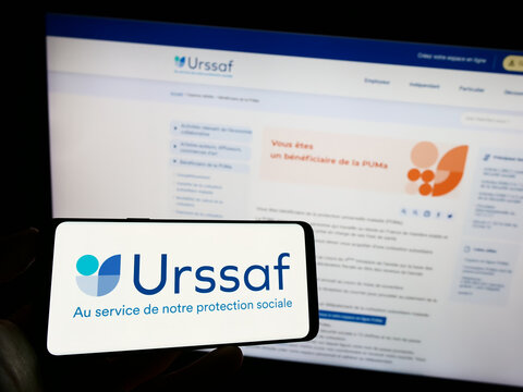 Stuttgart, Germany - 07-12-2023: Person holding mobile phone with logo of French social security organization URSSAF on screen in front of web page. Focus on phone display.