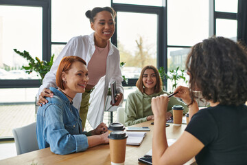 joyful african american coach with notebook embracing woman and talking to multicultural group sitting near takeaway drinks in interest club, mutual support and understanding concept