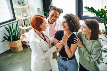 redhead motivation coach and smiling multicultural female friends embracing overjoyed multiracial...