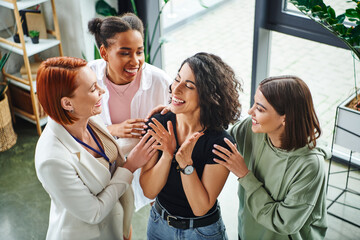high angle view of happy motivation coach and multicultural girlfriends embracing happy multiracial woman smiling and gesturing with closed eyes, moral support and mental wellness concept