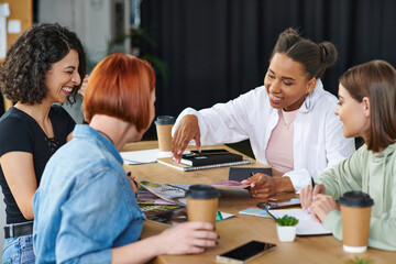 joyful african american woman pointing at magazines while spending happy time with multicultural girlfriends near coffee to go in women club, common interests and knowledge-sharing concept