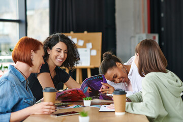 excited multicultural female friends reading beauty and style magazine and laughing near paper cups with takeaway drinks on table in women club, common interests and knowledge-sharing concept