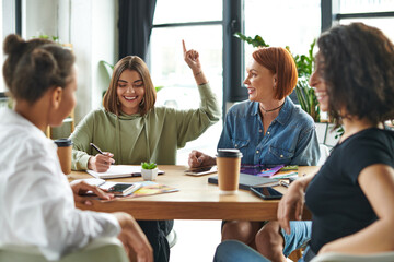 cheerful young woman showing idea gesture and writing in notebook near multiethnic friends and table with paper cups and magazines in female interest club, knowledge-sharing and socializing concept