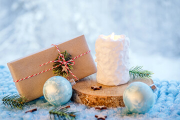 Christmas products background. White snowball pattern candle burning, Christmas present wrapped in brown paper with white and red string on wooden disk with bokeh snowy winter forest background.