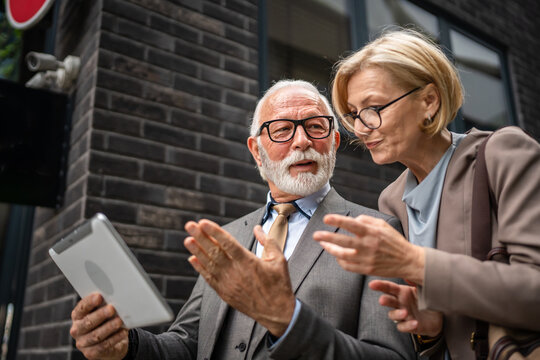 senior man dressed in a suit and a mature blonde woman as business partners husband and wife friends or colleagues talk With digital tablet in hand browse internet look data exchange ideas copy space