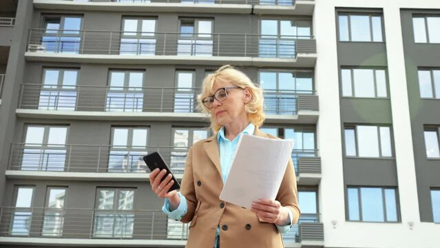 Female employee real estate agent writes information on smartphone from clipboard against building. Working process and negotiations with client low angle shot