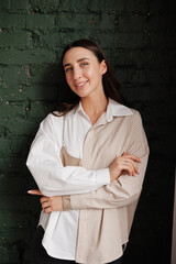 Attractive confidence young stylish woman in trendy shirt looks at camera, smiles, stands with crossed arms on brick wall. Beauty, fashion, self-expression, creativity, individuality concept