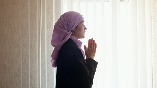 Muslim woman in pink hijab praying for better peaceful life in semi-dark apartment. Religious woman hopes Allah for best opportunities in life