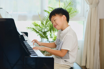 Cute happy smiling little Asian kid boy playing piano in living room at home, Elementary school child having fun with learning to play music instrument, Music education concept