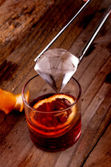 putting diamond ice into red negroni drink with campari and vermouth on wooden table with orange...