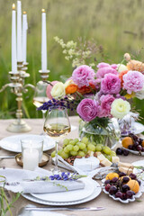 Elegant fancy wedding table decor with bright ranunculi flowers in a vase outdoors, chandelier, field or meadow. Summer, sunset, golden hour. Romantic dinner for a couple in restaurant outside
