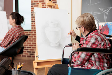 Senior woman wheelchair user drawing with pencil on canvas, attending art classes for people with disability, reducing stress in creative space. Creative therapy for disabled adults