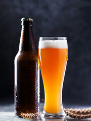 A glass of wheat beer and a cold beer bottle in drops of water, wheat ears. dark background
