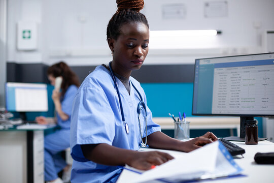 African american nurse inside modern, clean, clinical office, sitting at desk. Young professional adult woman employee focused on comparing data between computer and documents