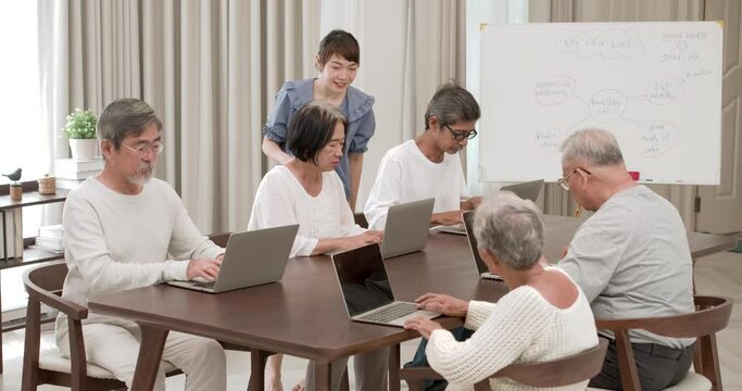 A beautiful kindness young Asian nurse walking around and teaching seniors male and female to using computer in classroom. Teacher take care of older men and women to learn about modern technology.