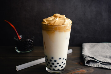 Iced Dalgona Coffee with Boba in a Pint Glass on a Dark Background: Cold dalgona coffee with milk and tapioca pearls shown with a boba straw