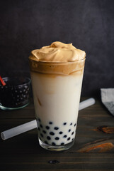 Iced Dalgona Coffee with Boba in a Pint Glass on a Dark Background: Cold dalgona coffee with milk and tapioca pearls shown with a boba straw