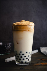 Iced Dalgona Coffee with Boba in a Pint Glass on a Dark Background: Cold dalgona coffee with milk and tapioca pearls shown with a boba straw