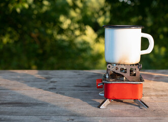 White enamel cup on a portable camping gas burner outdoors, copy space. Camping or picnic concept