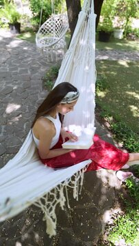 The beautiful hippie girl is swinging in a hammock and flipping through a book.