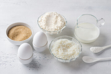 Ingredients for cooking white chocolate coconut muffins: flour, milk or buttermilk, eggs, coconut flakes, brown sugar, salt and baking powder on a light gray table