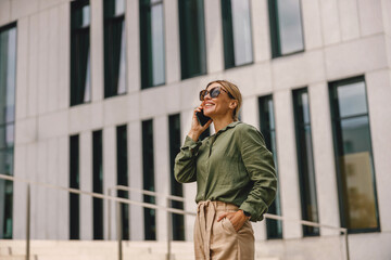 Stylish woman entrepreneur talking by phone whiile standing on modern building background 