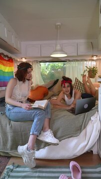 Two beautiful hippie girls are on the bed inside the trailer, with a laptop and a book.