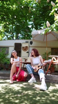 slow motion of Two beautiful hippie girls are singing a song sitting on the terrace near the trailer with a guitar.