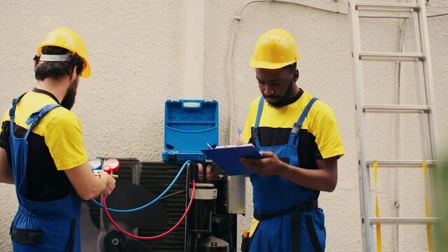 Expert technicians team working with manifold gauges to check air conditioner refrigerant levels, writing result on clipboard. Skillful mechanics using barometer benchmarking hvac system tool