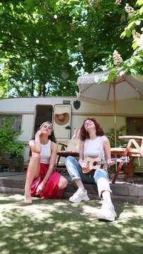 Two beautiful hippie girls are singing a song sitting on the terrace near the trailer with a guitar.