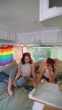 Two beautiful hippie girls are talking on the bed inside the trailer with a laptop.