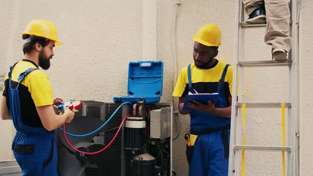 Capable electrician team working with manometers to check air conditioner freon levels, writing result on clipboard. Seasoned wiremen using barometer benchmarking condenser tool