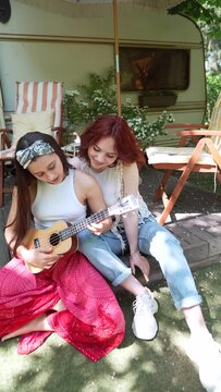 Two beautiful hippie girls are singing a song sitting on the terrace near the trailer with a guitar.