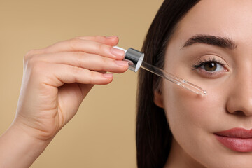 Young woman applying essential oil onto face on beige background, closeup