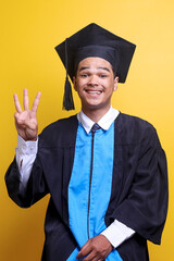 Young Asian man wearing graduation cap and ceremony robe with happy face showing number three and looking at the camera
