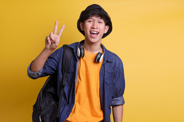 Cheerful male college student showing two fingers v-sign peace greeting gesture in casual clothes, headphones around his neck and wear a bucket hat 