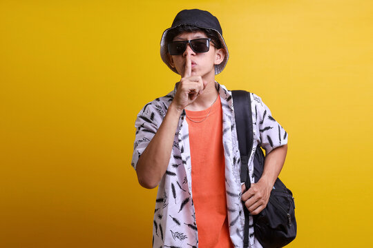 Trendy young man wearing sunglasses, bucket hat and carrying backpack put his index finger to his lips make silence, ssh or be quiet gesture.