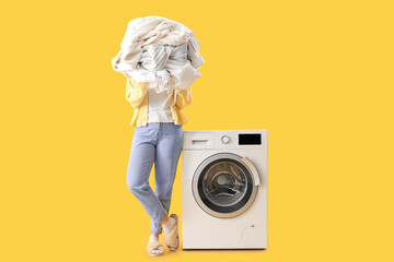 Woman holding pile of dirty clothes near washing machine on yellow background