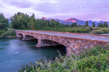 A stone bridge spanning a wide river with trees mountain in the background in the soft light of pre...