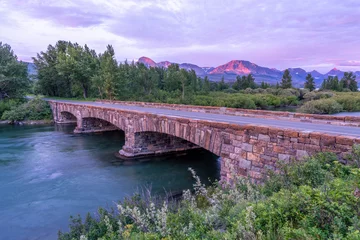 Fototapeten A stone bridge spanning a wide river with trees mountain in the background in the soft light of pre sunrise, Saint Mary Lake Bridge, Glacier National Park, Montana © Ron