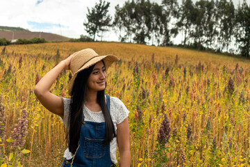 mujer joven latina con sombrero sonriendo de perfil en frente de un campo de Quinoa amarilla...