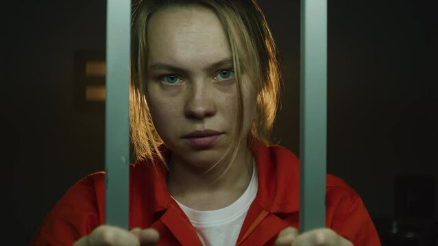 Female prisoner in orange uniform shakes, holds metal bars, looks at camera, then goes to window in prison cell. Woman criminal serves imprisonment term in jail or correctional facility. Portrait.