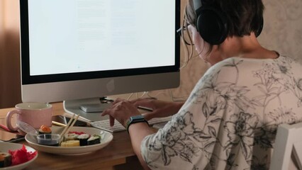 Adult female freelancer hunched over, sitting finishing urgent project, typing on keyboard in front monitor with white screen, plates of half-eaten Japanese rolls lying on desktop in home office.