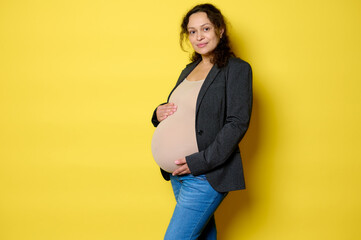 Attractive Latina pregnant woman in stylish blazer, bodysuit and blue jeans, smiles at camera,...